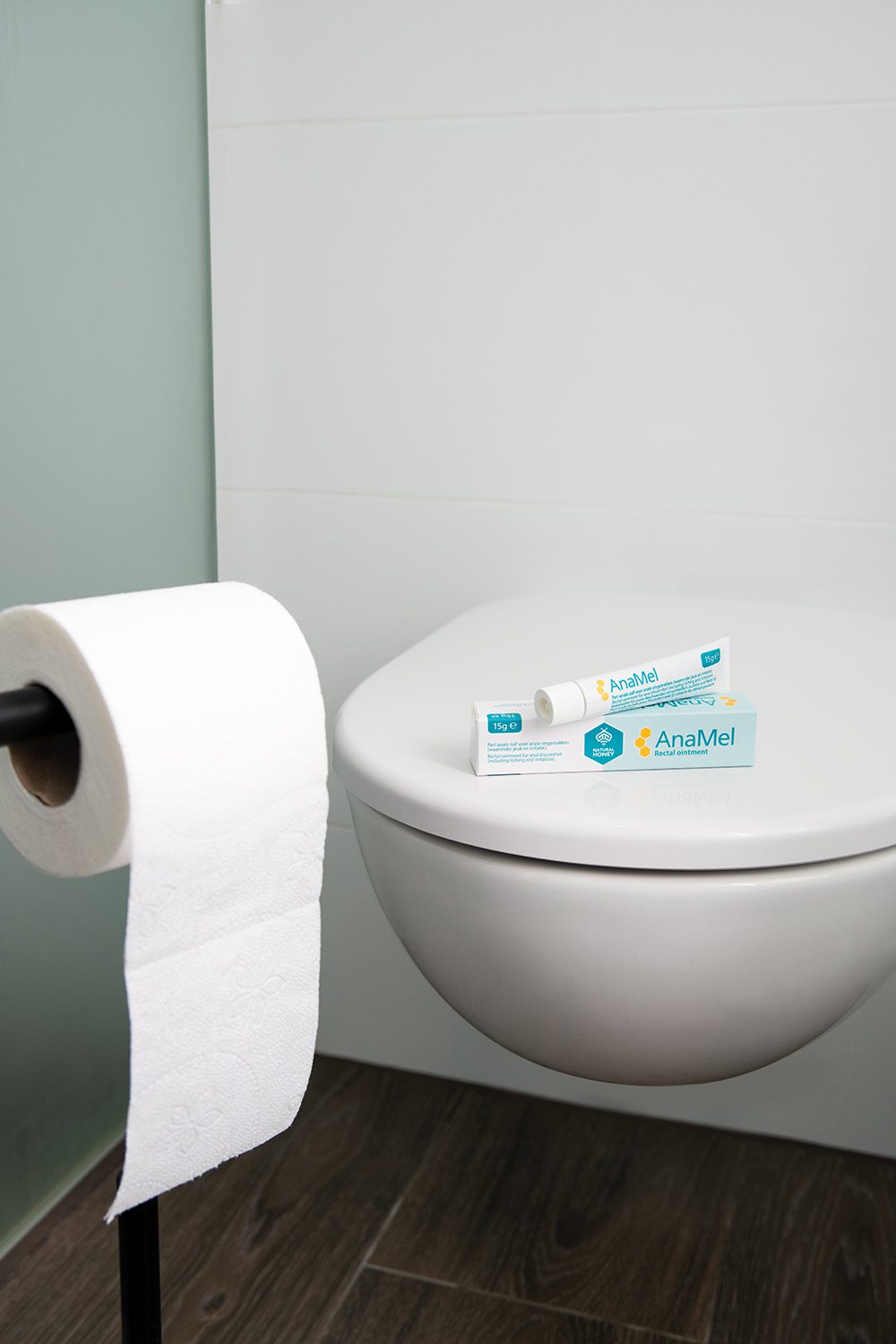 Toilet with a box of AnaMel on top, next to a roll of toilet paper.